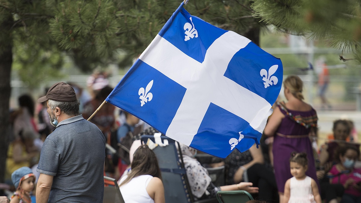 Un homme portant un masque tient un grand drapeau du Québec dans un parc. Derrière lui, des enfants et des femmes sont assises.