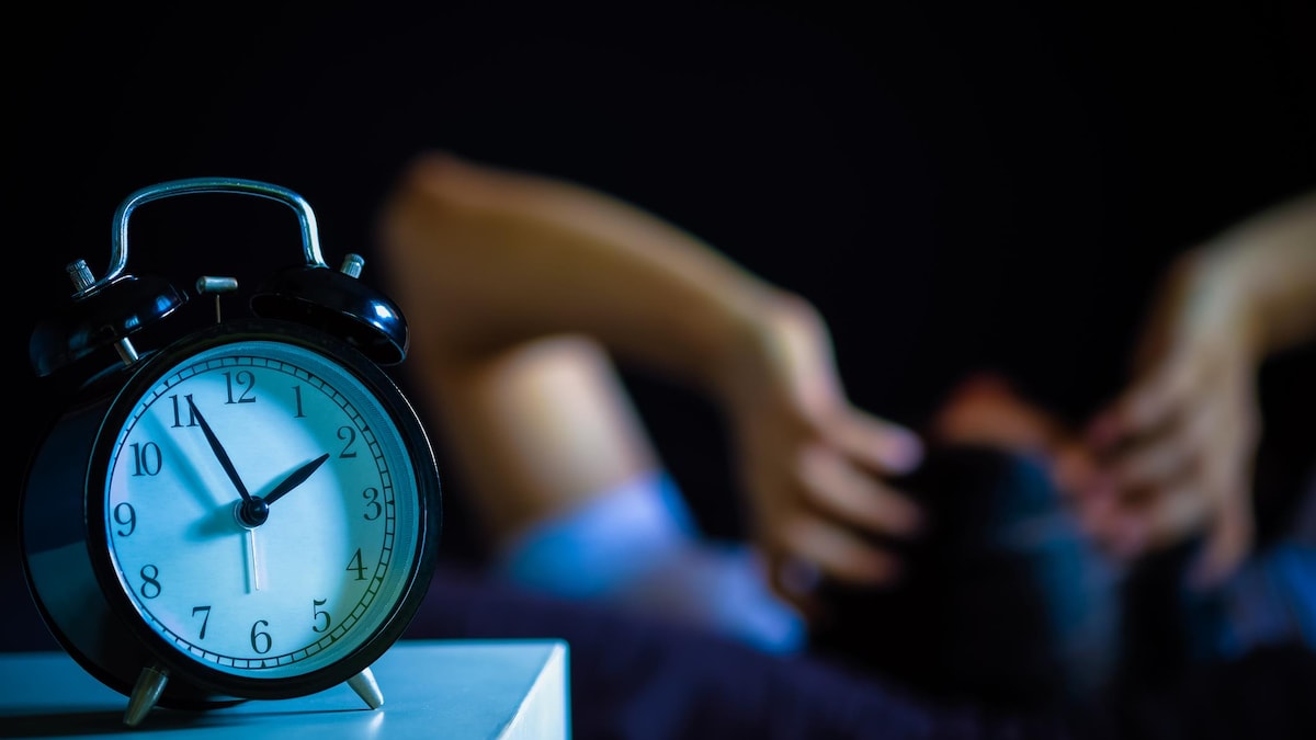 Une personne allongée se tient la tête dans le fond de l'image avec une horloge au premier plan qui indique 1 h 55.