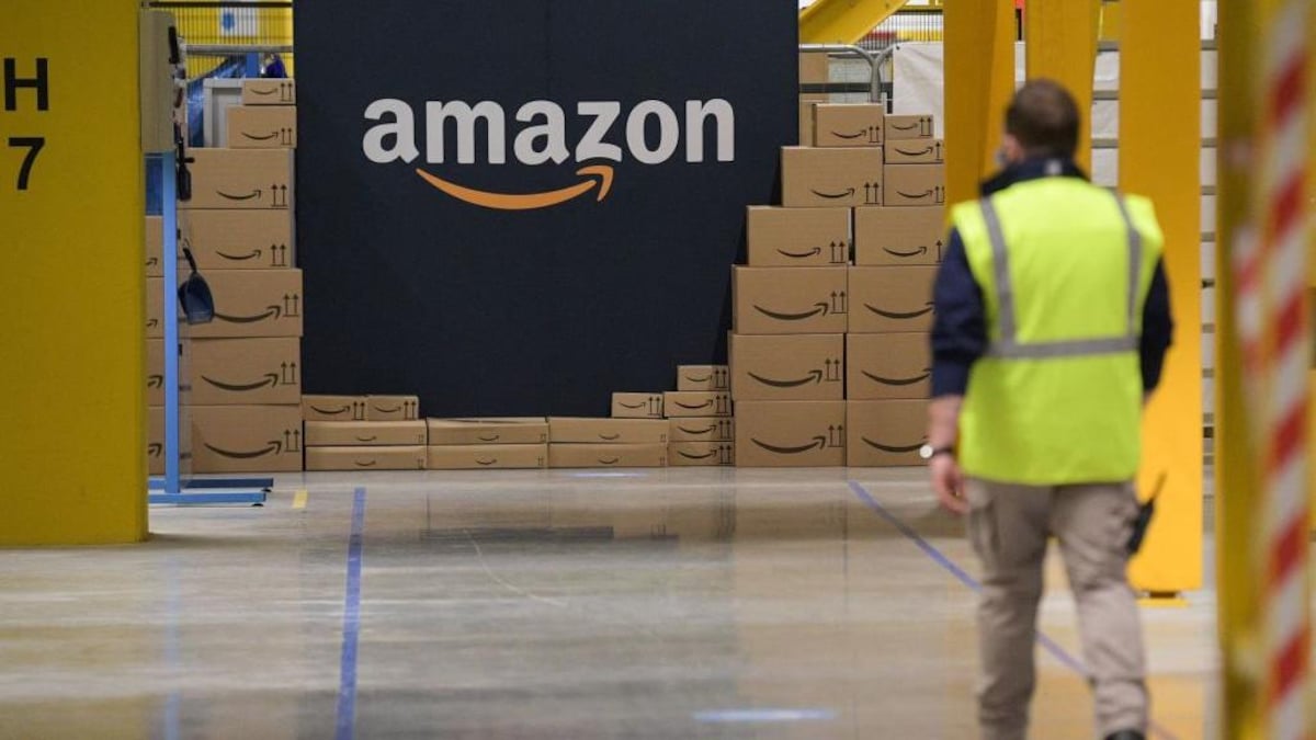 Une personne avec un gilet de sécurité marche dans un hangar avec une grande pancarte amazon dans le fond entourée de cartons de la même entreprise.